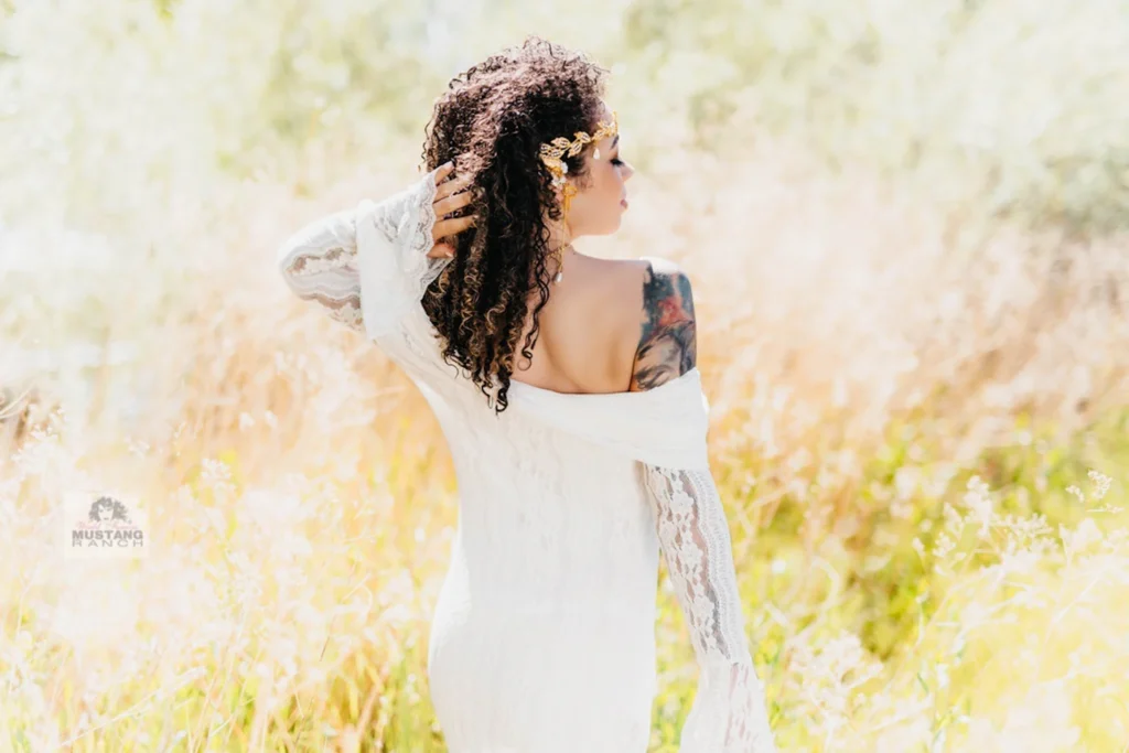 Side-profile of a woman in a white lace dress in a sunlit field, with curly hair and a flower crown.