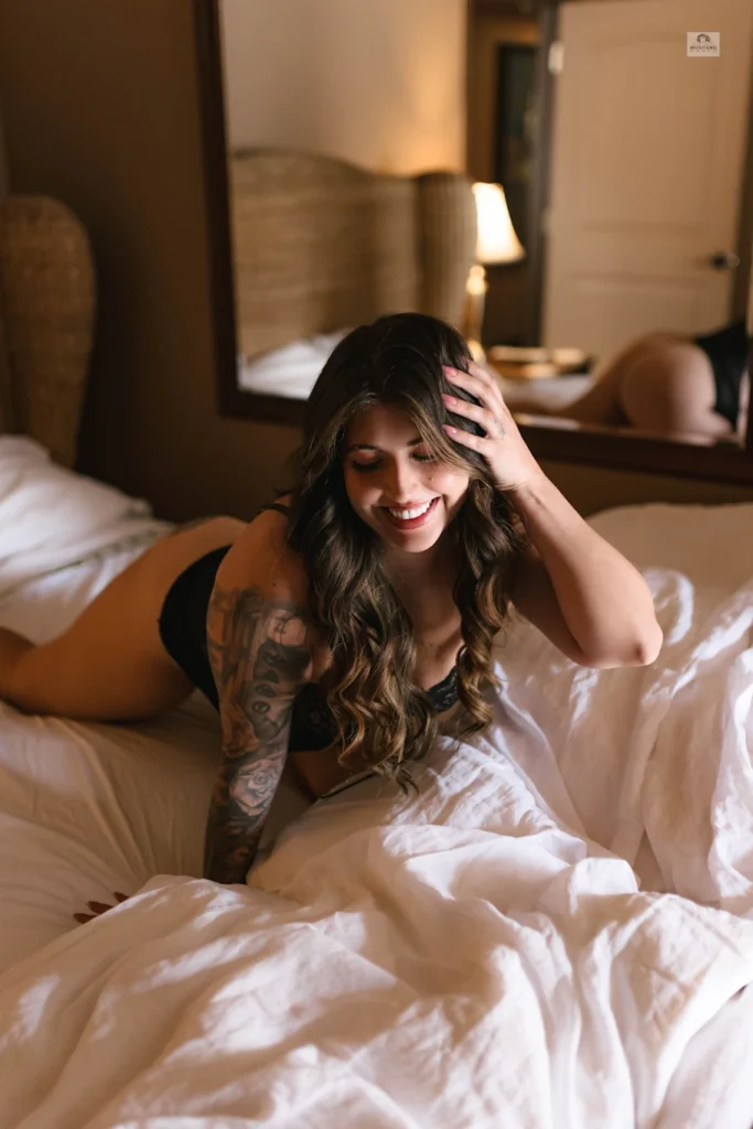 Woman in black lingerie on a bed, smiling and brushing hair with one hand.