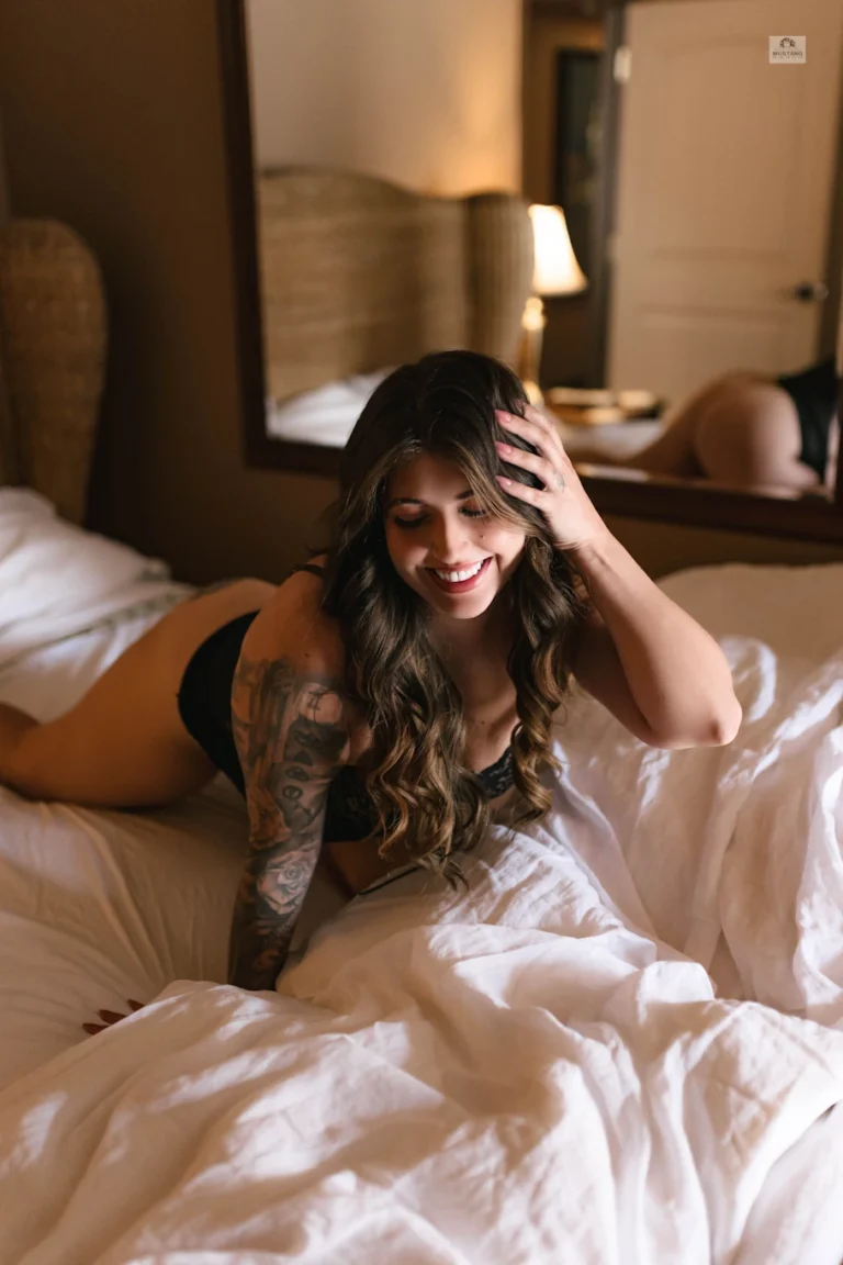 Woman in black lingerie on a bed, smiling and brushing hair with one hand.