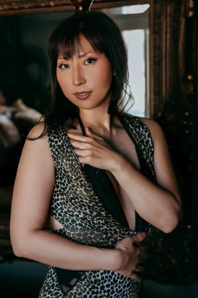 Woman wearing a leopard-print dress with a plunging neckline, one hand on her chest, posing in a softly lit room.
